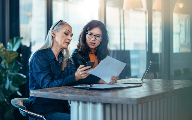 Two business woman sitting at a table and working together. Senior executive showing her phone and papers to young colleague at office.